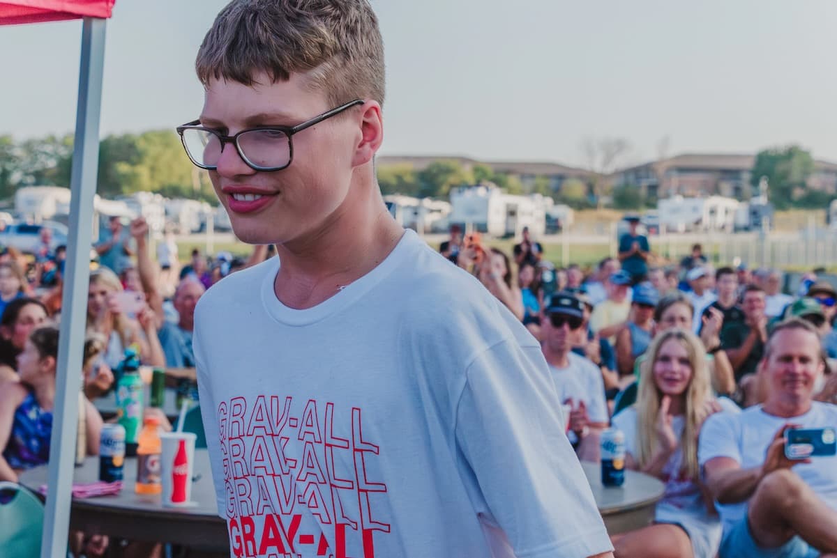 Young athlete in blue Adventure for All shirt smiling at outdoor Gravel Worlds race venue with spectators visible in background