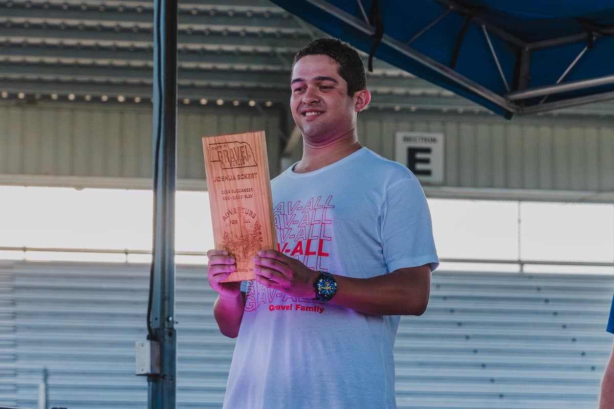 Athlete holding personalized cherry wood number plate holder trophy for Joshua Eckert, 2024 Buccaneer 50K, 2,000 ELEV race category, with "Adventure for All" branding and landscape engraving