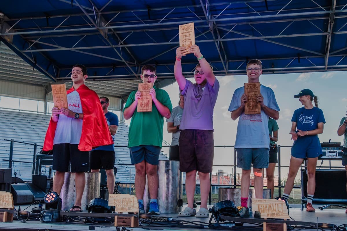 Five young athletes on stage at Gravel Worlds 2024 race awards ceremony holding CNC-carved cherry wood trophy plaques with Adventure for All branding and personalized engraving, under blue tent structure