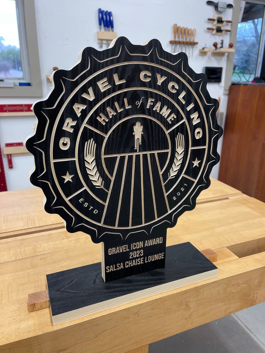 Gravel Cycling Hall of Fame trophy plaque for Salsa Chaise Lounge, Class of 2023. Black ash wood with gold-engraved star badge design and cyclist figure, displayed on light wood workshop table.