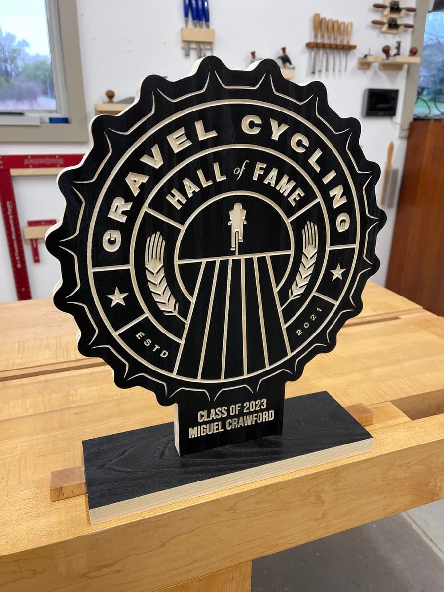 Gravel Cycling Hall of Fame trophy plaque for Miguel Crawford, Class of 2023. Black ash wood with gold-engraved star badge design and cyclist figure, displayed on light wood workshop table.