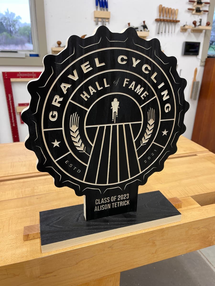 Gravel Cycling Hall of Fame trophy plaque for Alison Tetrick, Class of 2023. Black ash wood with gold-engraved star badge design and cyclist figure, displayed on light wood workshop table.
