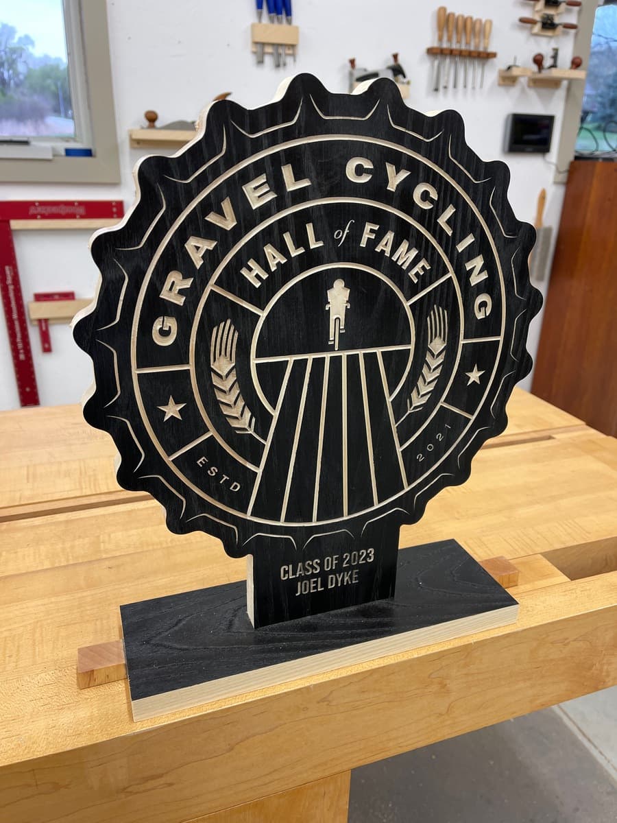 Gravel Cycling Hall of Fame trophy plaque for Joel Dyke, Class of 2023. Black ash wood with gold-engraved star badge design and cyclist figure, displayed on light wood workshop table.