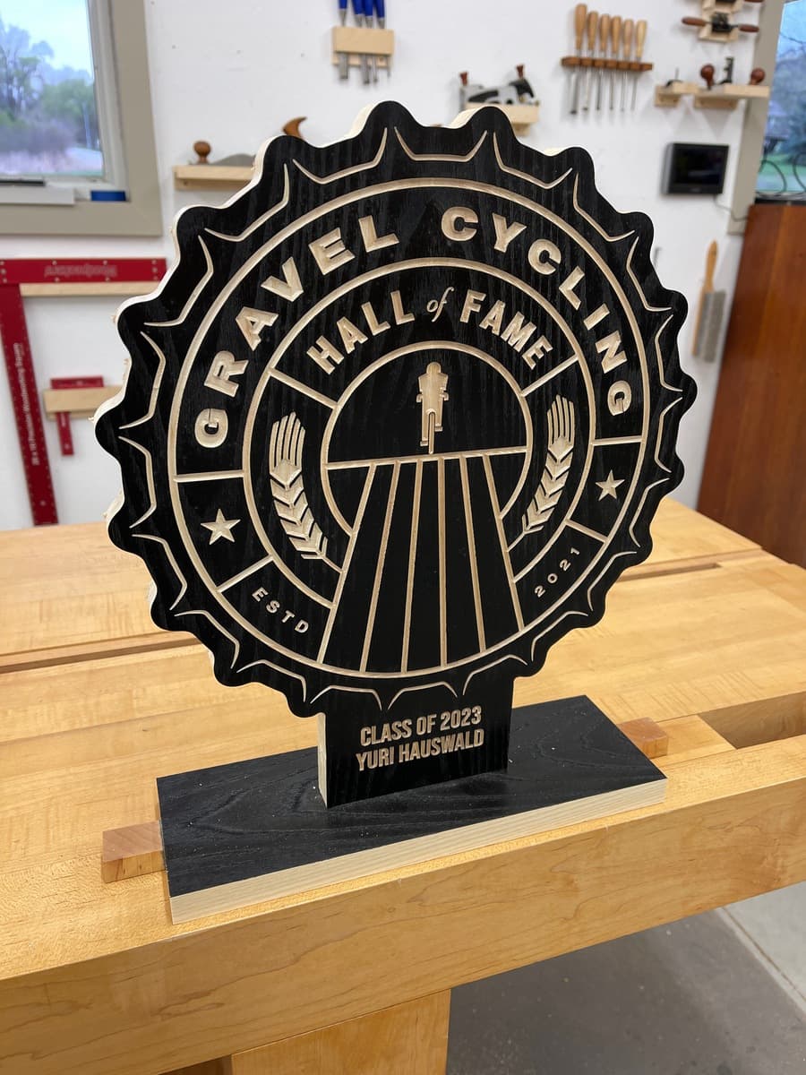 Gravel Cycling Hall of Fame trophy plaque for Yuri Hauswald, Class of 2023. Black ash wood with gold-engraved star badge design and cyclist figure, displayed on light wood workshop table.