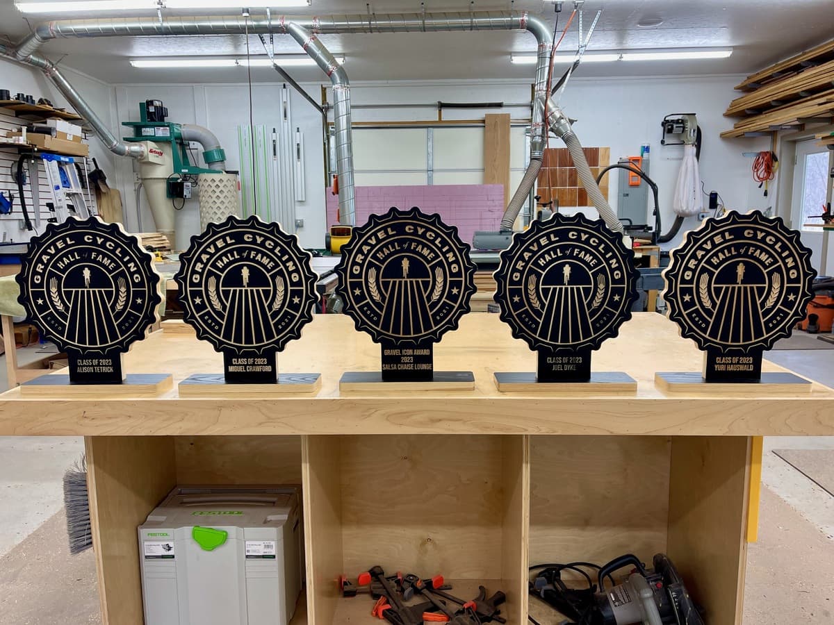 Five completed Gravel Cycling Hall of Fame trophy plaques in a row on workshop table. Black ash wood bases with gold-engraved vintage badge designs and cyclist figures for Class of 2023 winners.