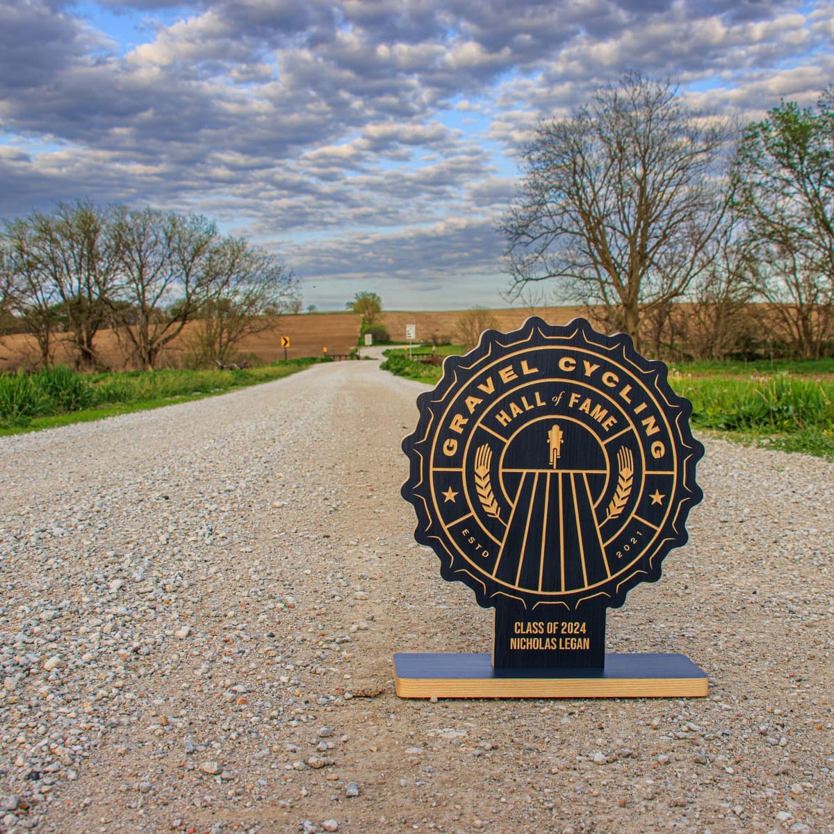 Four completed Gravel Cycling Hall of Fame trophies displayed in a row on rural gravel driveway. Navy blue ash wood bases with gold-engraved badge designs for Class of 2024 winners: Jay Peterbary, Nicholas Legan, Linda Guerrette, and Selene Yeager.