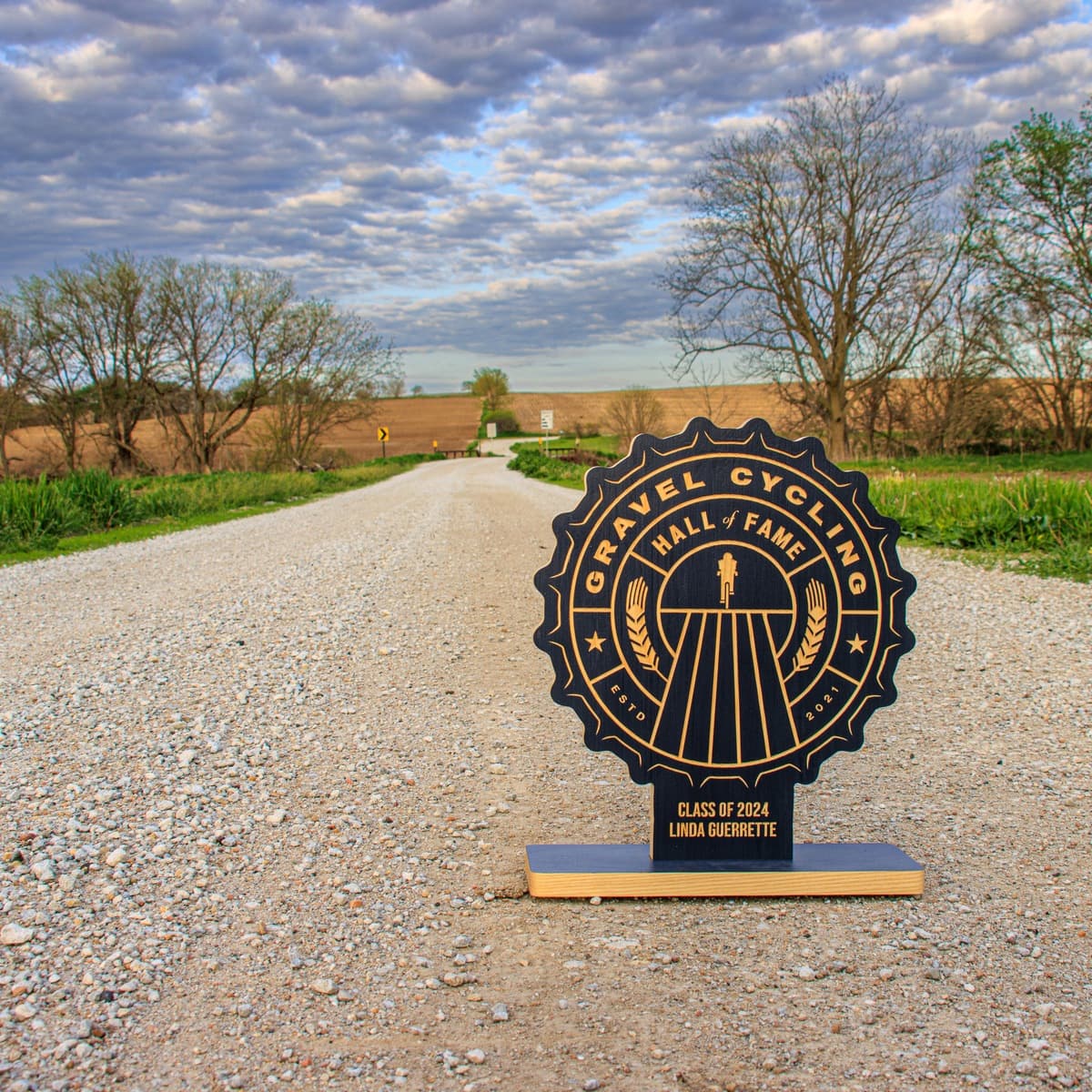 Gravel Cycling Hall of Fame trophy for Nicholas Legan, Class of 2024. Navy blue ash wood base with gold-engraved custom design featuring cyclist figure, grain motifs, and vintage badge aesthetic on rural gravel road background.