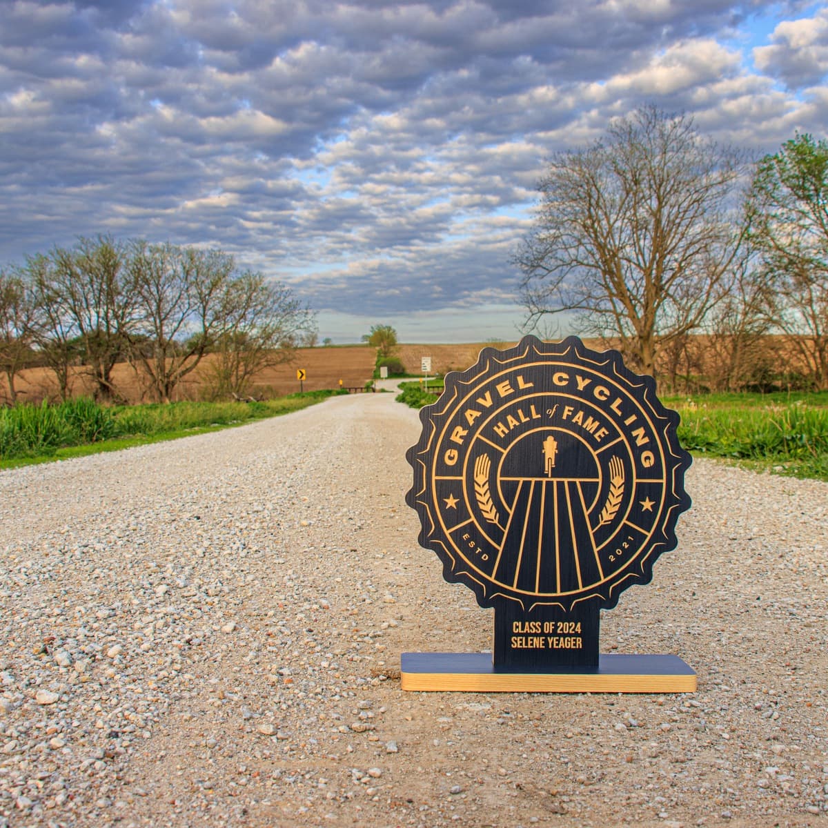 Gravel Cycling Hall of Fame trophy for Linda Guerrette, Class of 2024. Navy blue ash wood base with gold-engraved custom design featuring cyclist figure, grain motifs, and vintage badge aesthetic on rural gravel road background.