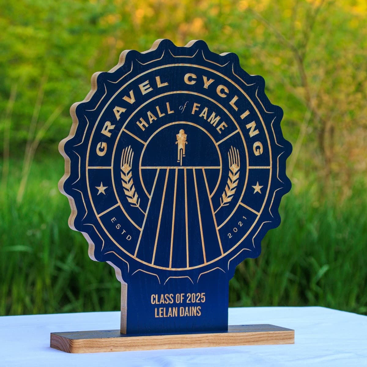 Gravel Cycling Hall of Fame trophy plaque for Lelan Dains, Class of 2025. Navy blue ash wood base with gold-engraved custom design featuring cyclist figure and vintage badge aesthetic.