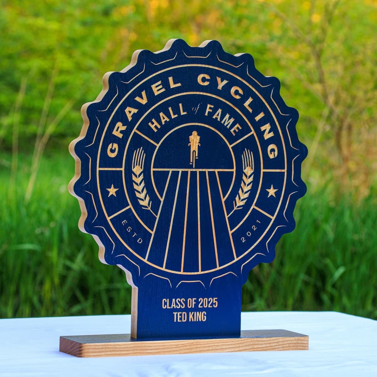 Gravel Cycling Hall of Fame trophy plaque for Ted King, Class of 2025. Navy blue ash wood base with gold-engraved custom design featuring cyclist figure and vintage badge aesthetic.