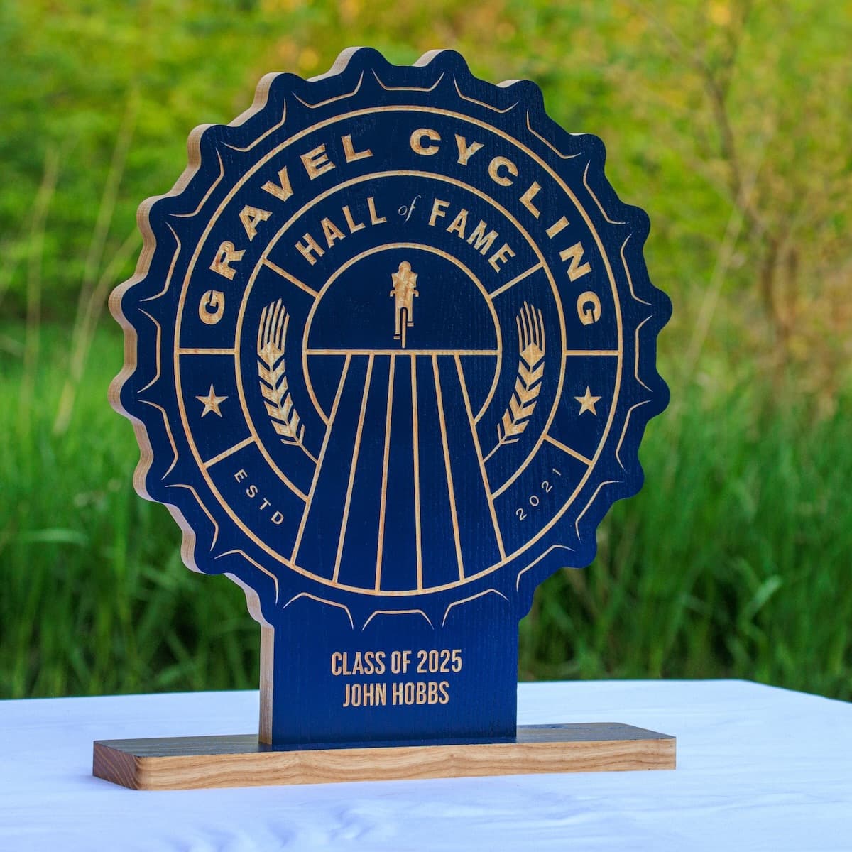 Gravel Cycling Hall of Fame trophy plaque for John Hobbs, Class of 2025. Navy blue ash wood base with gold-engraved custom design featuring cyclist figure and vintage badge aesthetic.