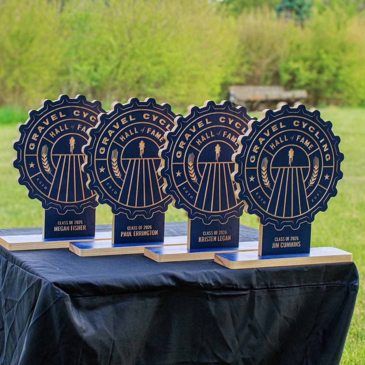 Four Gravel Cycling Hall of Fame Class of 2026 inductee trophies displayed on a table with black tablecloth and green outdoor background. Navy blue ash gear-shaped plaques for Megan Fisher, Paul Errington, Kristen Legan, and Jim Cummins.