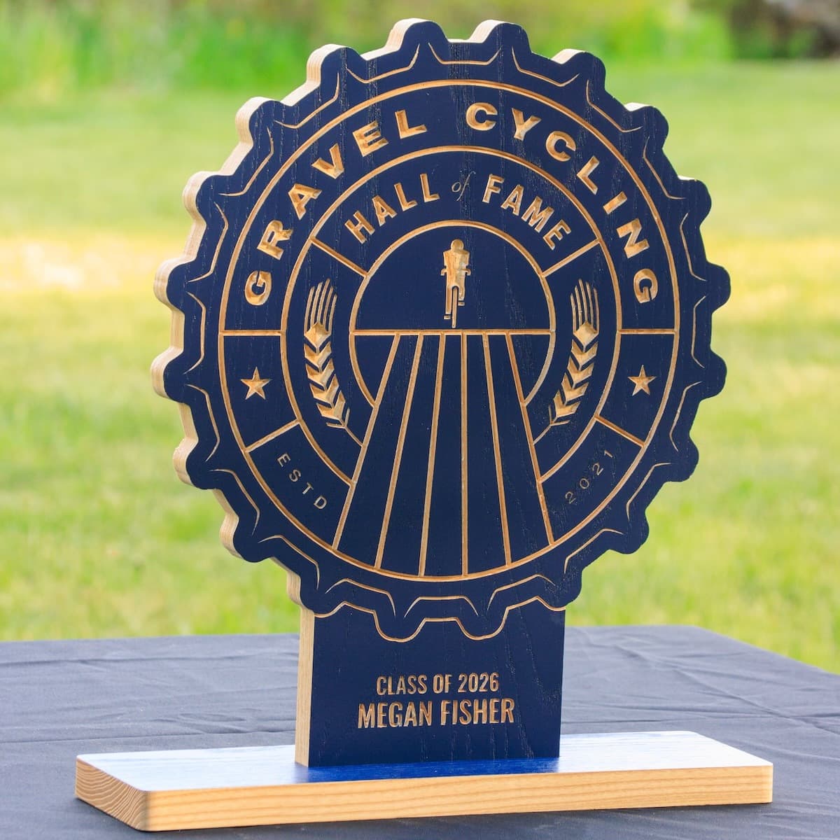 Gravel Cycling Hall of Fame trophy for Megan Fisher, Class of 2026. Navy blue ash wood gear-shaped plaque with CNC-carved cyclist silhouette on gravel road and natural ash base.