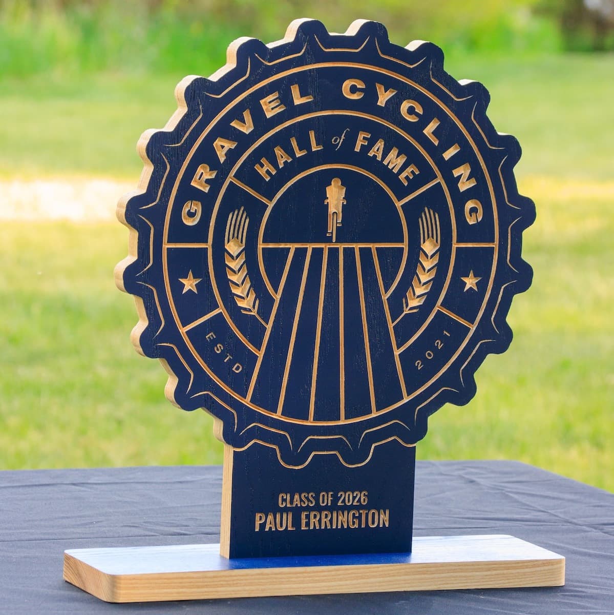 Gravel Cycling Hall of Fame trophy for Paul Errington, Class of 2026. Navy blue ash wood gear-shaped plaque with CNC-carved gravel cycling design and natural ash base.