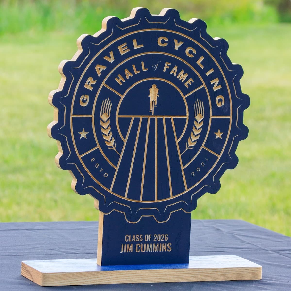 Gravel Cycling Hall of Fame trophy for Jim Cummins, Class of 2026. Navy blue ash wood gear-shaped plaque with CNC-carved cyclist silhouette design and natural ash base.