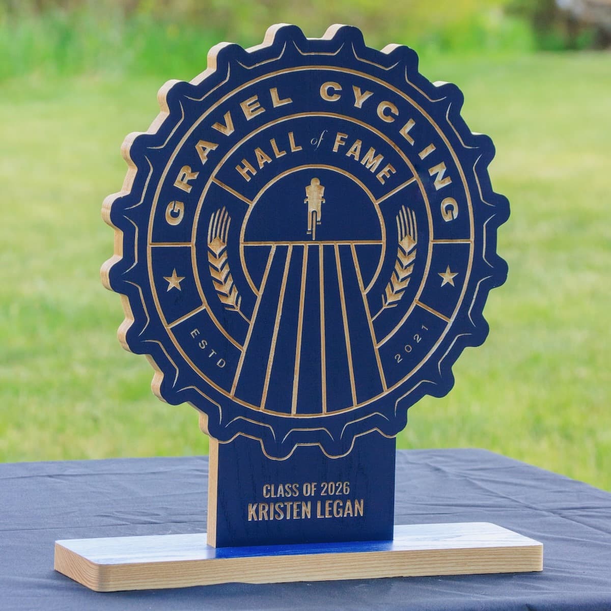 Gravel Cycling Hall of Fame trophy for Kristen Legan, Class of 2026. Navy blue ash wood gear-shaped plaque with CNC-carved cyclist silhouette and natural ash base.