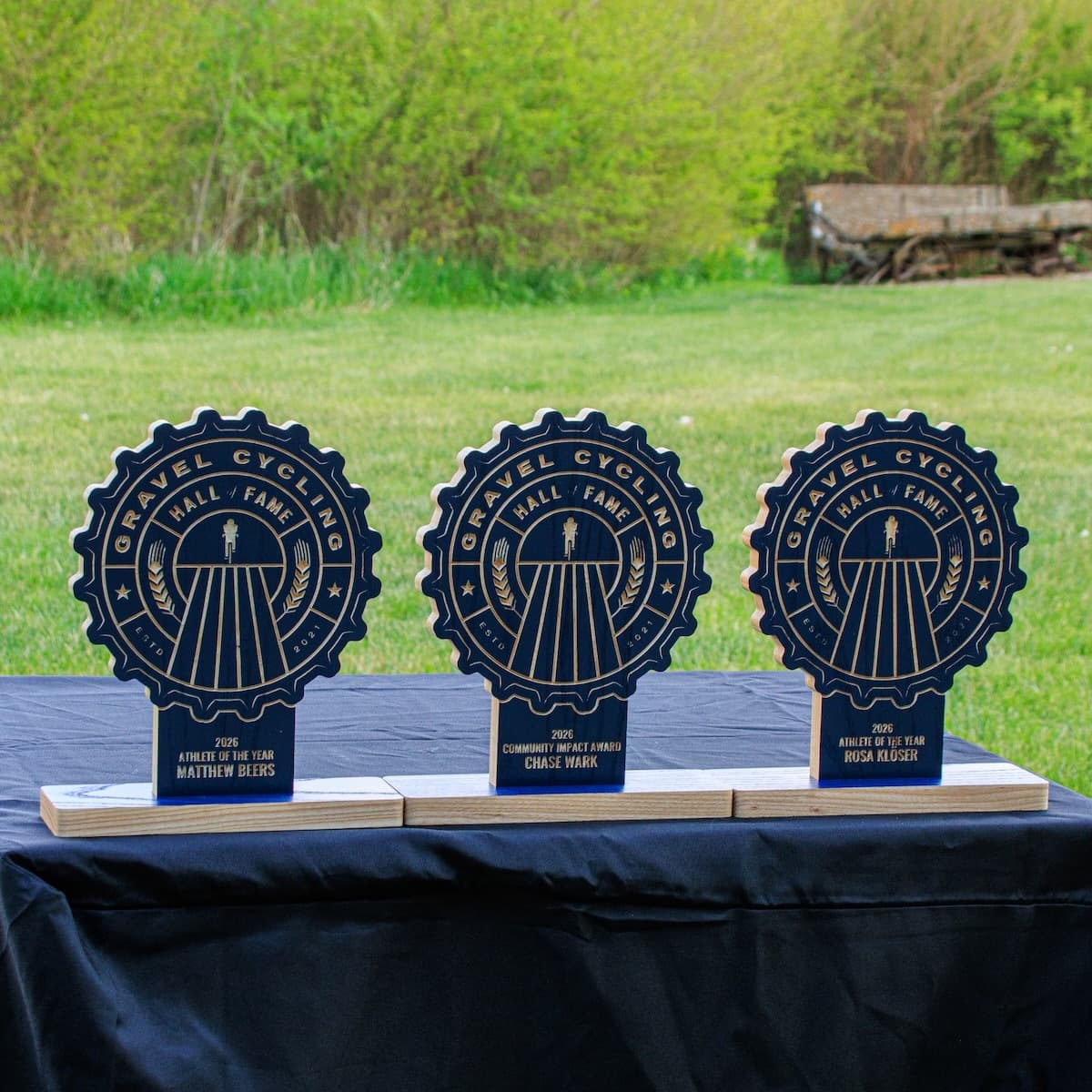 Three Gravel Cycling Hall of Fame 2026 award trophies on black tablecloth outdoors. Left to right: Matthew Beers (Athlete of the Year), Chase Wark (Community Impact Award), Rosa Klöser (Athlete of the Year). Navy blue ash gear-shaped design.