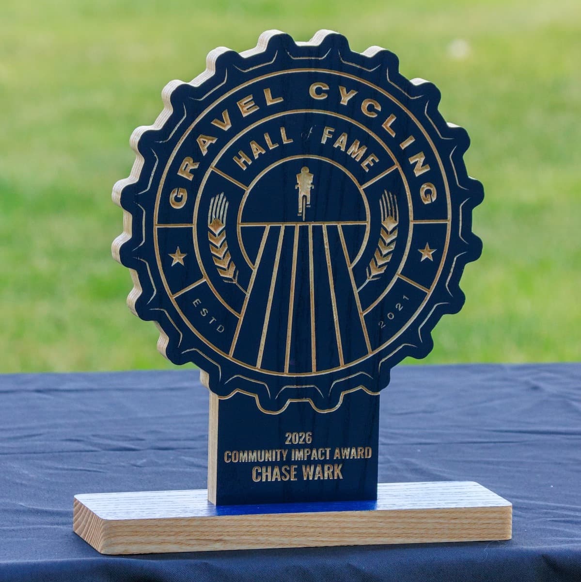 Gravel Cycling Hall of Fame 2026 Community Impact Award trophy for Chase Wark. Navy blue ash wood gear-shaped plaque with CNC-carved gravel cycling design and natural ash base with blue accent.