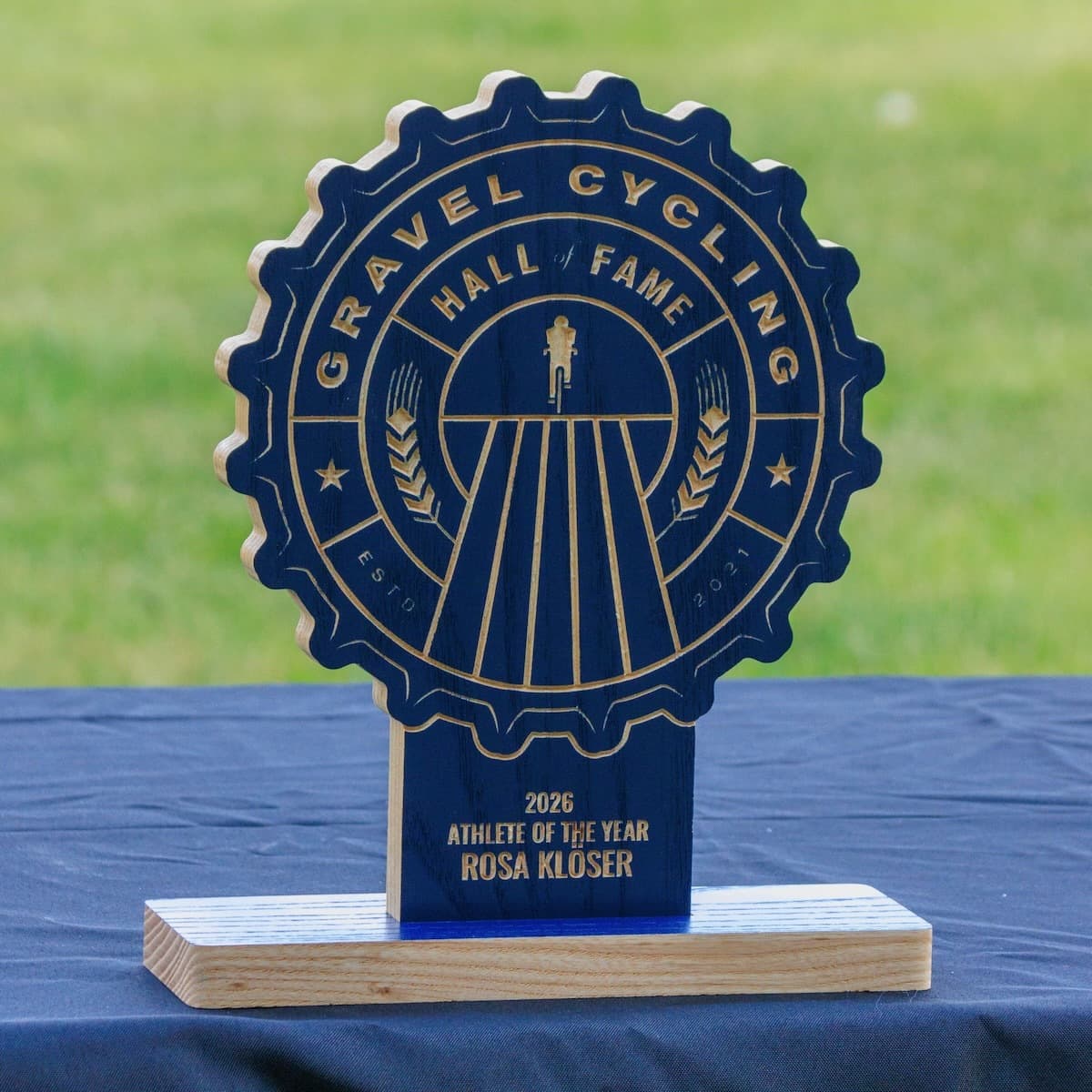 Gravel Cycling Hall of Fame 2026 Athlete of the Year trophy for Rosa Klöser. Navy blue ash wood gear-shaped plaque with CNC-carved cyclist silhouette on gravel road and natural ash base with blue accent.