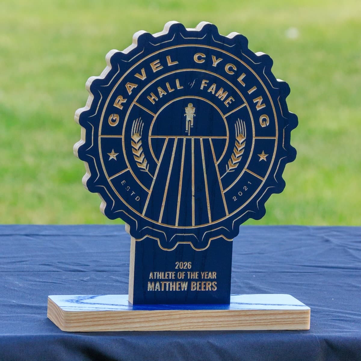 Gravel Cycling Hall of Fame 2026 Athlete of the Year trophy for Matthew Beers. Navy blue ash wood gear-shaped plaque with CNC-carved design and natural ash base with blue accent.