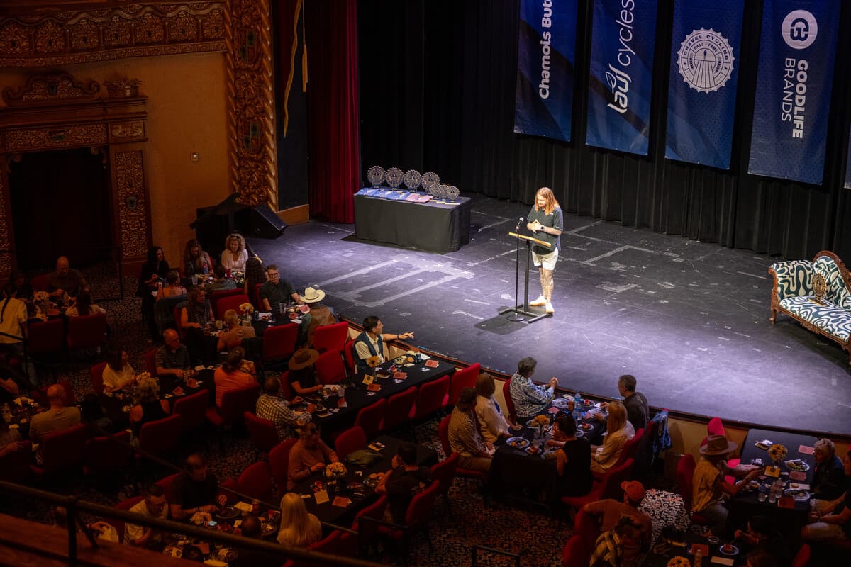 Wide overhead view of the Gravel Cycling Hall of Fame 2026 induction ceremony at a historic theater. A presenter stands at a podium on stage with all the ash wood trophies displayed on the table behind her. Guests are seated at round tables in the audience. Sponsor banners including Gravel Cycling Hall of Fame, Chamois Butt'r, and GoodLife Brands hang from the stage.