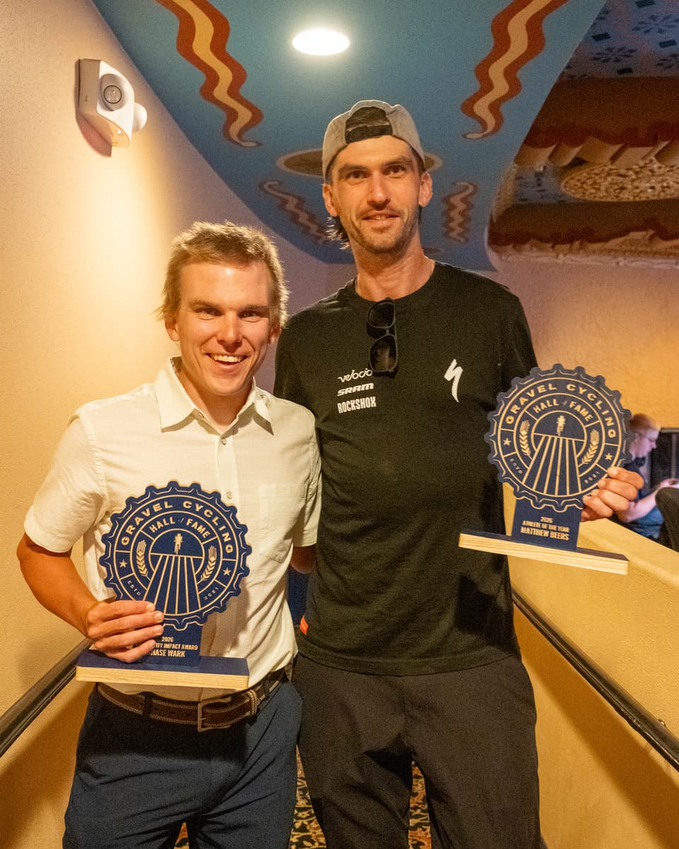 Chase Wark (Community Impact Award) and Matthew Beers (Athlete of the Year) pose together in a hallway at the GCHOF 2026 ceremony, both smiling and holding their navy blue ash wood Gravel Cycling Hall of Fame trophies.