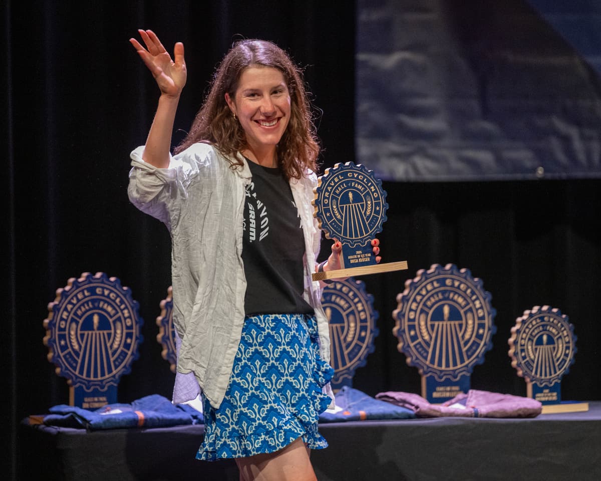 Rosa Klöser waves to the crowd on stage at the GCHOF 2026 ceremony while holding her Athlete of the Year trophy. Additional inductee trophies are displayed on the stage table in the background.