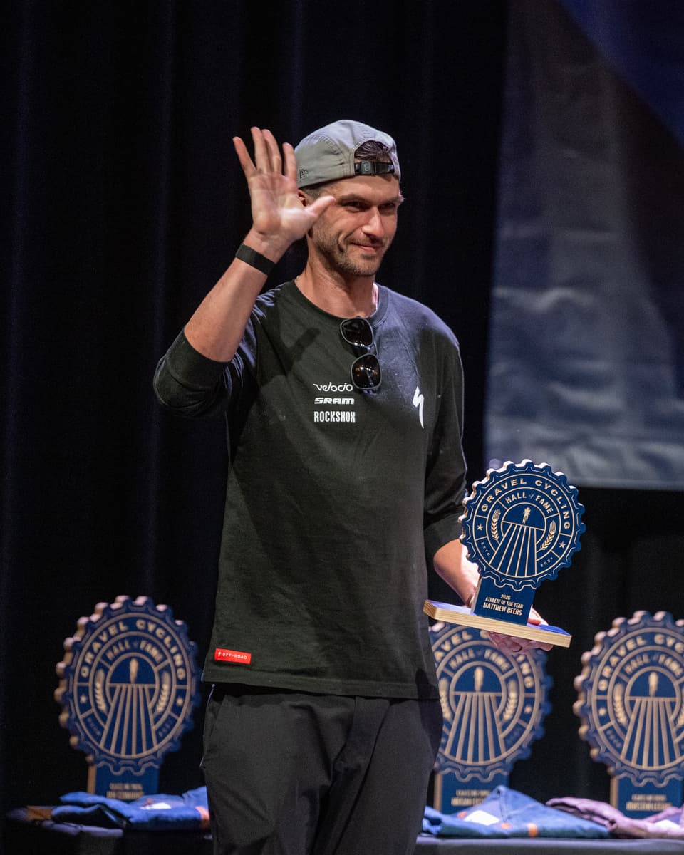 Matthew Beers waves to the audience on stage at the GCHOF 2026 ceremony, holding his Athlete of the Year trophy. Additional trophies are visible on the stage table in the background.