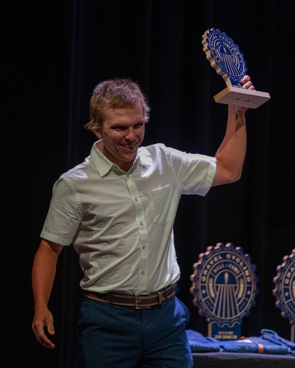 Chase Wark raises his Gravel Cycling Hall of Fame Community Impact Award trophy overhead in celebration on stage at the GCHOF 2026 ceremony. Other inductee trophies are visible on the table behind him.