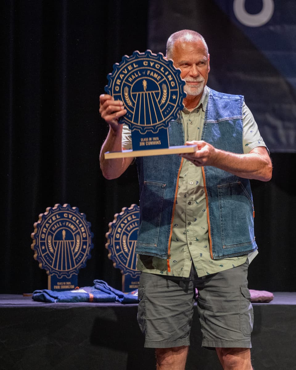Jim Cummins holds up his Gravel Cycling Hall of Fame Class of 2026 inductee trophy on stage at the ceremony, displaying the navy blue ash gear-shaped plaque with his name engraved on the base.