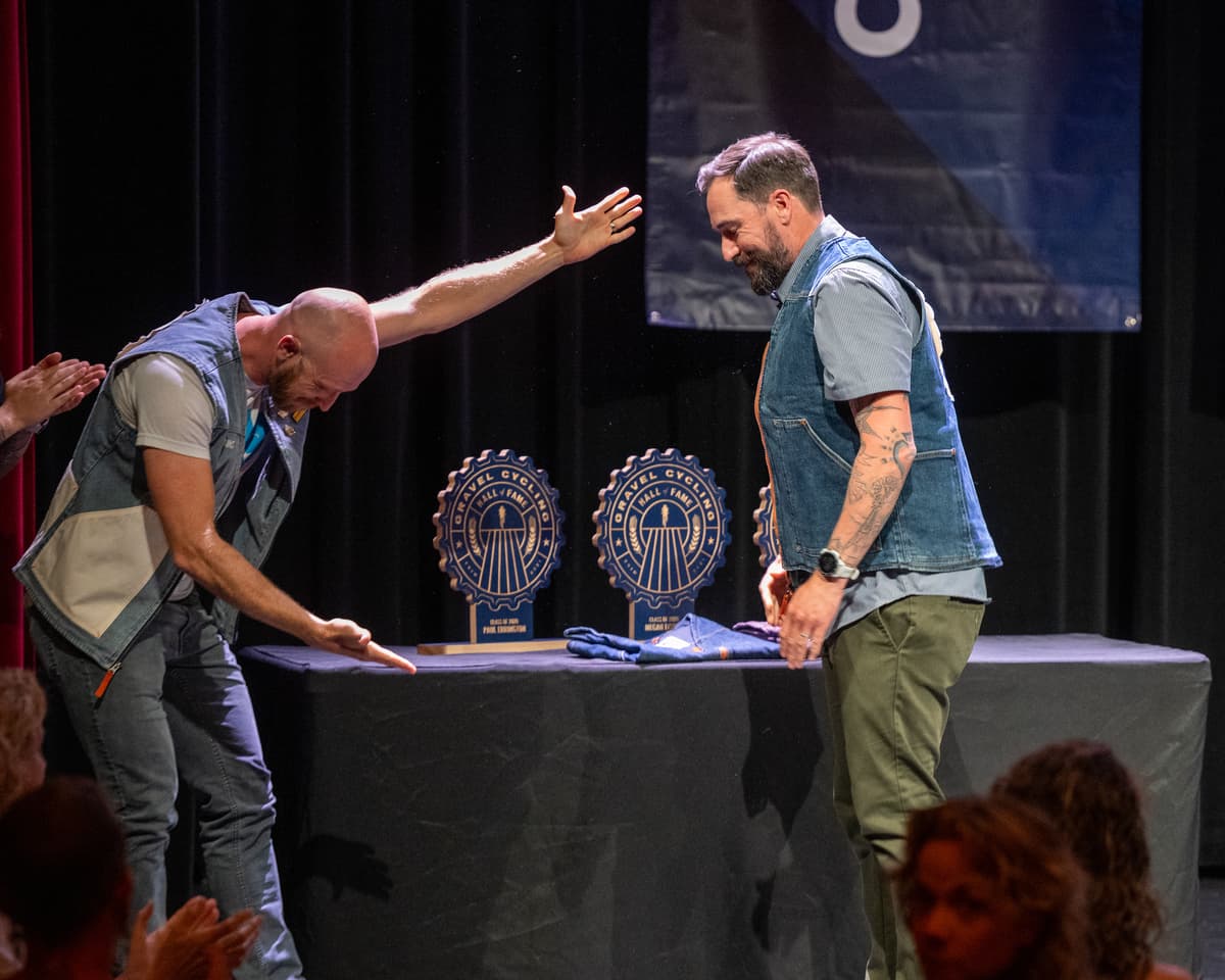 Paul Errington (left) enthusiastically gestures at the trophy table while Jim Cummins (right) laughs beside him on stage at the GCHOF 2026 ceremony. Two ash wood trophies for Paul Errington and Megan Fisher sit on the table between them.