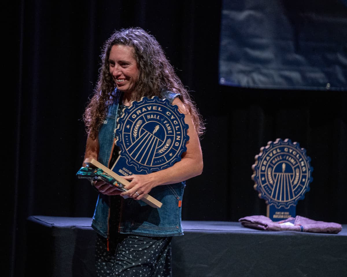 Megan Fisher smiles as she holds her Gravel Cycling Hall of Fame Class of 2026 inductee trophy on stage at the ceremony. Kristen Legan's trophy is visible in the background on the stage.