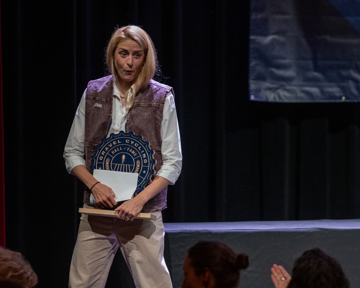 Kristen Legan walks across the stage at the GCHOF 2026 ceremony with a look of delighted surprise, holding her Hall of Fame inductee trophy. Audience members are visible in the foreground.