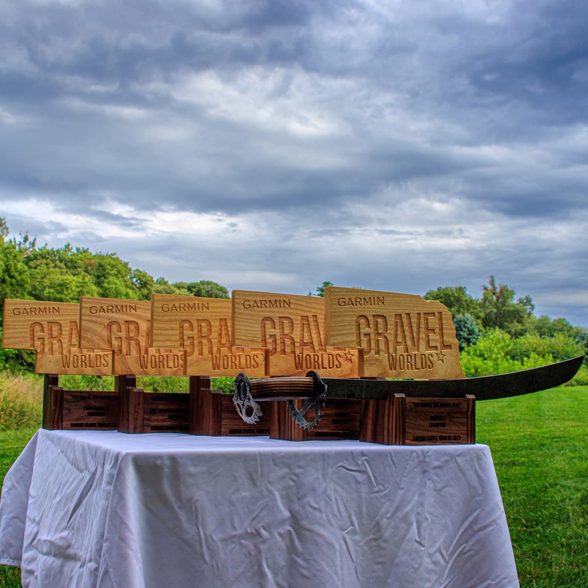Five wooden Garmin Gravel Worlds trophies with engraved text and sword graphics displayed on white tablecloth outdoors, showing progressive sizing against blue sky and trees