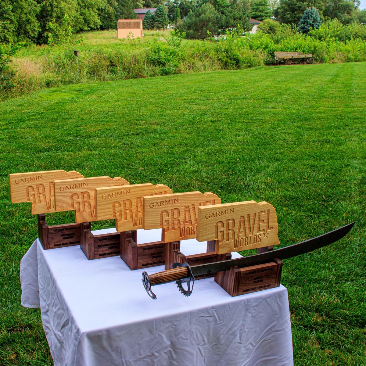 Five Gravel Worlds wooden sword holder trophies arranged in a row on white table displaying varied sizes and walnut/ash wood construction with engraved plaques