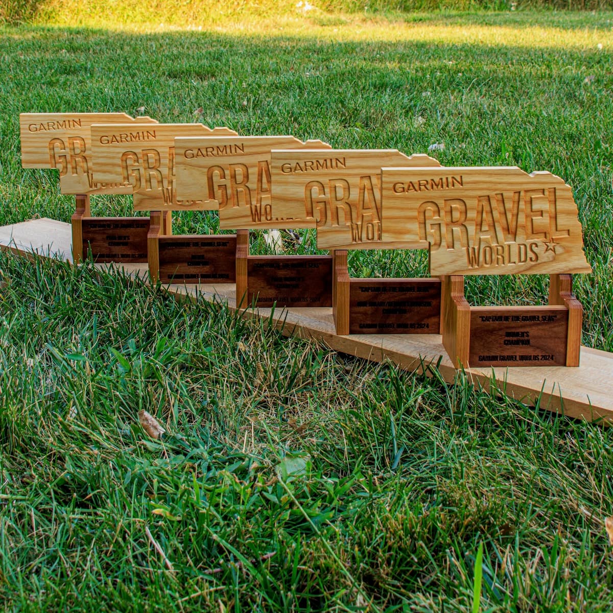 Five wooden Garmin Gravel Worlds trophies with engraved text displayed in grass, showing progressive sizing