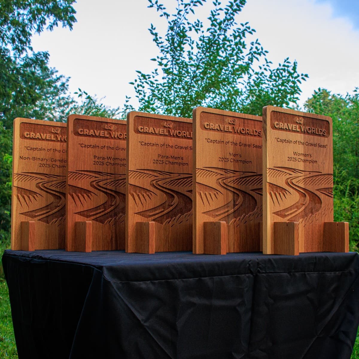 Five wooden sword holder trophies lined up outdoors with engraved Gravel Worlds branding and championship category text