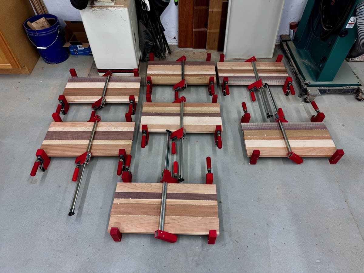 Workshop overhead view of numerous multi-species wood number plate holder plaques in progress, clamped with red clamps and metal bars for drying