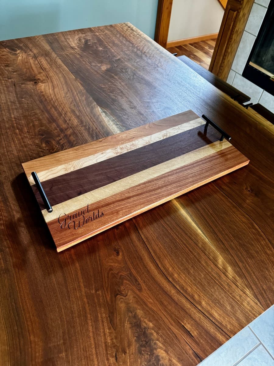 Single "Gravel Worlds" branded wooden number plate holder with black metal handle, showcasing engraved script text on walnut and light wood striped base