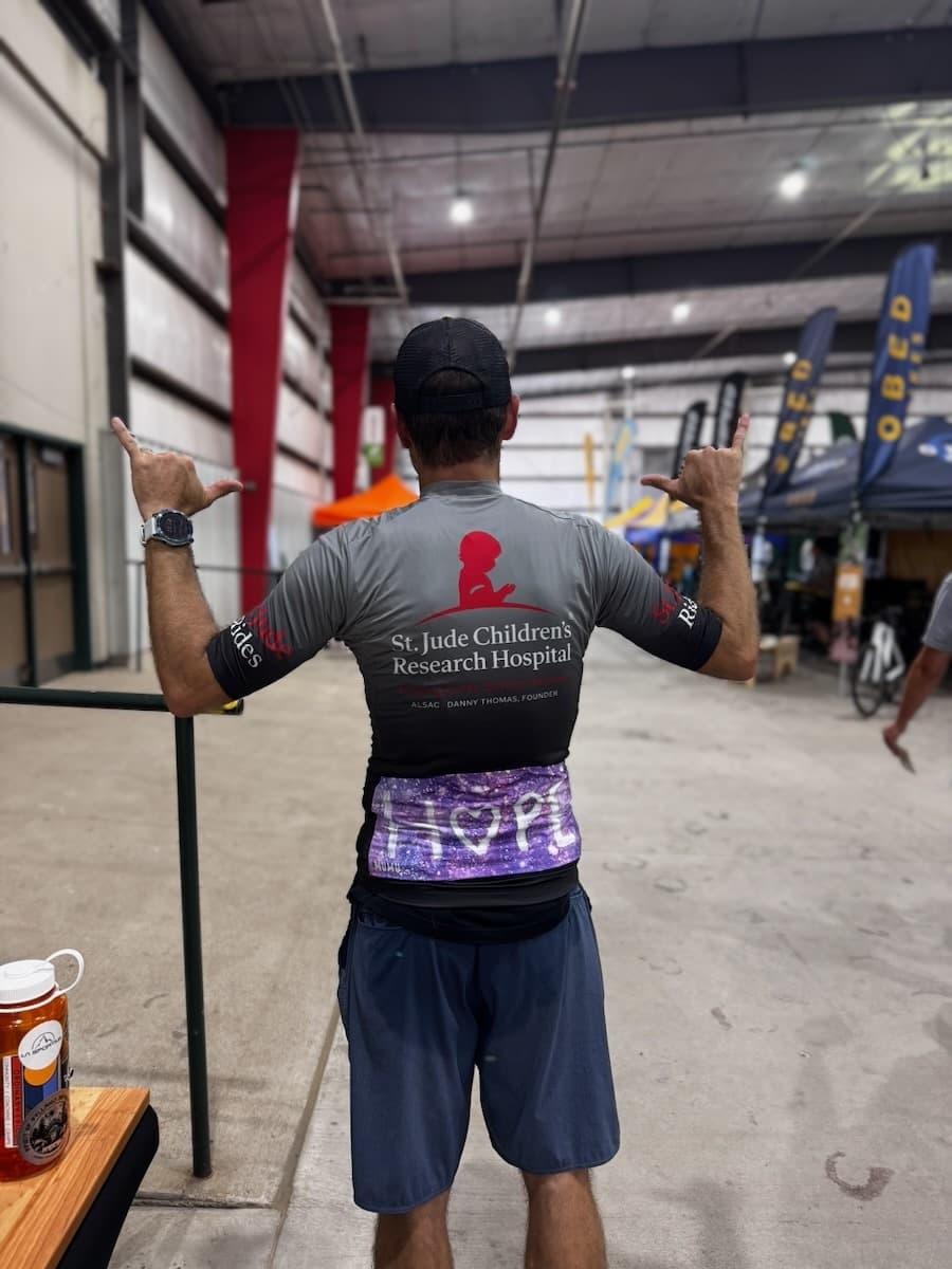 Exhibition booth display with wooden racing bib holder plaques and various merchandise arranged on a black table at Gravel Worlds event
