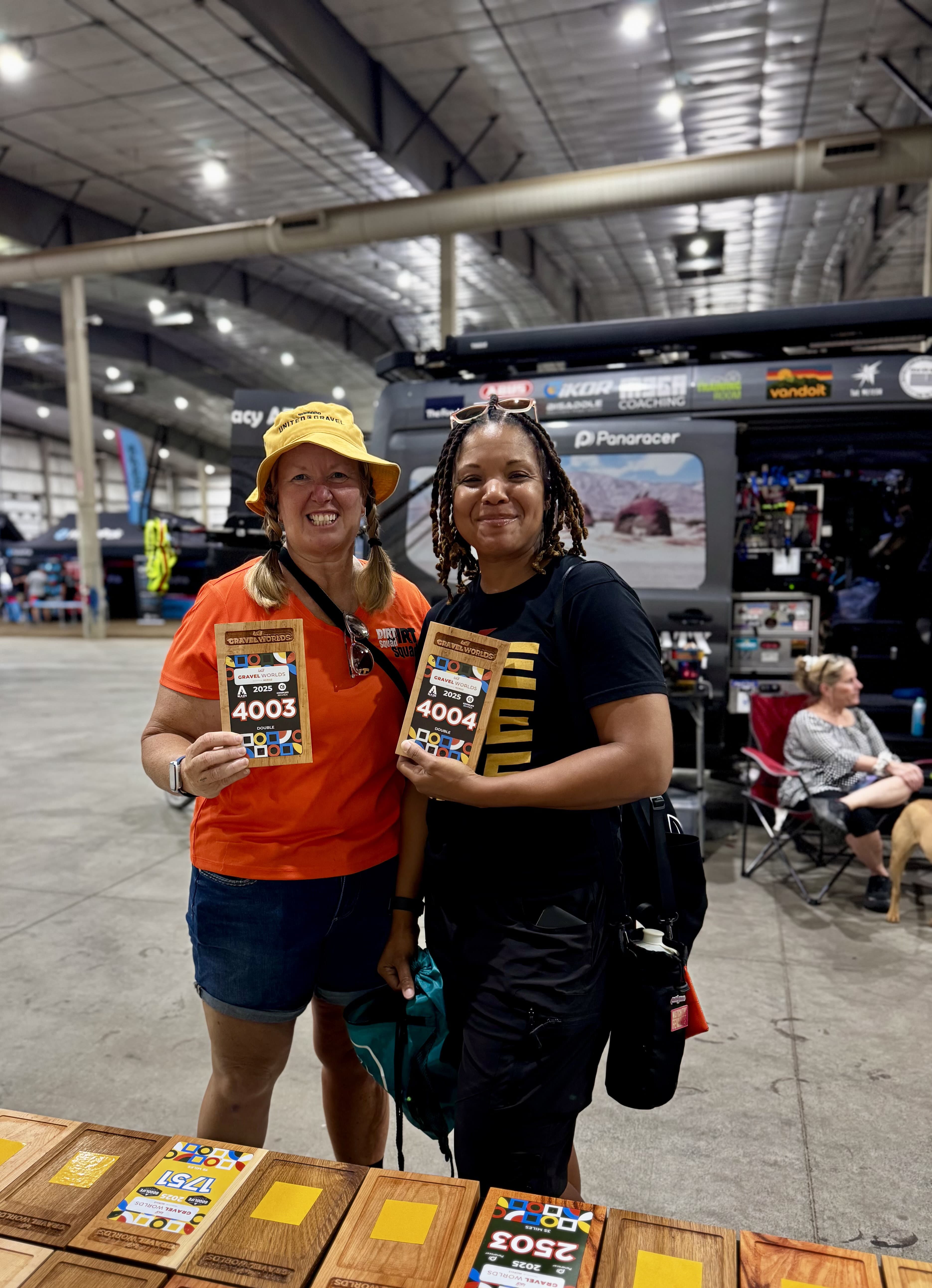 Mike posing with another person at the Gravel Worlds booth, both wearing St. Jude-related clothing, with wooden trophy plaques visible on display behind them