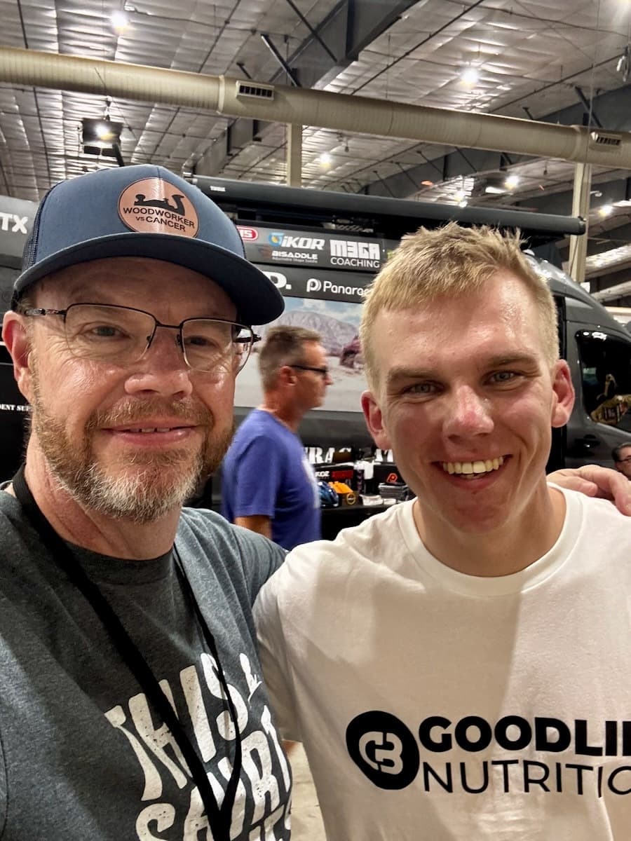 Mike in his Woodworker vs Cancer branded cap standing with another person at Gravel Worlds Expo event, with booth setup visible behind them