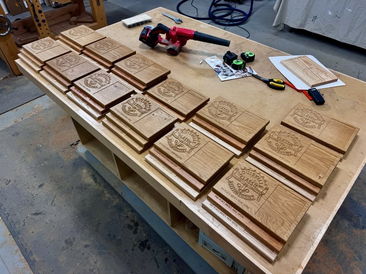Multiple CNC-engraved white oak trophy plaques arranged on workshop table with CNC cutting tools and supplies visible