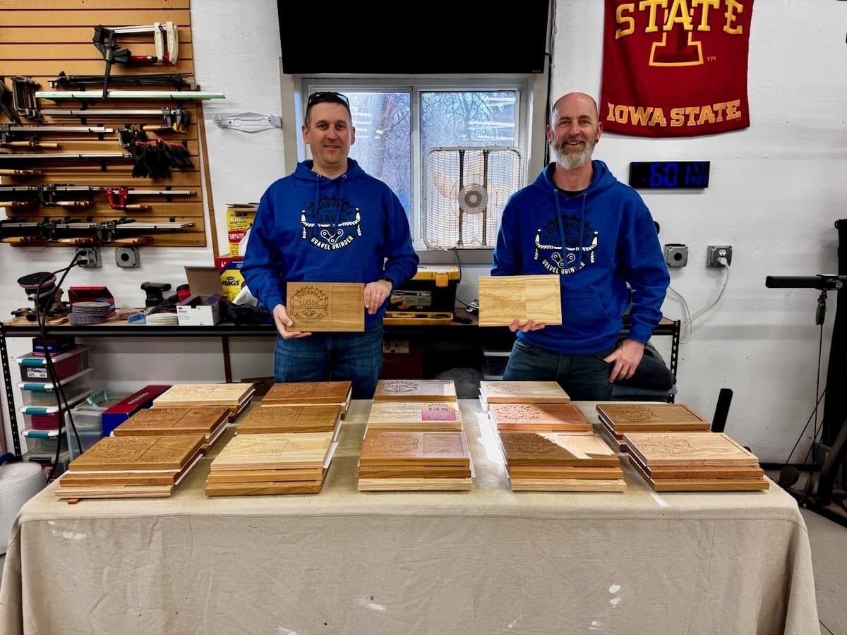 Two men in Louisville Classic Gravel Grinder hoodies holding finished white oak trophy plaques in workshop with shelving visible