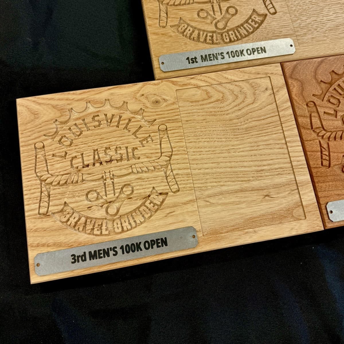 Multiple Louisville Classic Gravel Grinder trophy plaques arranged on dark background showing 3rd Men's 100K Open and other race categories with blue labels