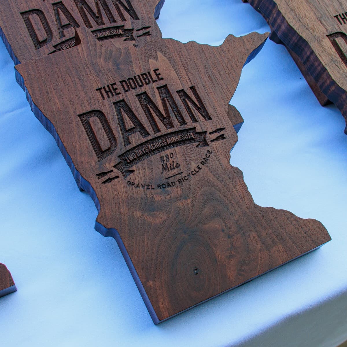 Wooden Minnesota-shaped trophy with CNC-engraved text reading "THE DOUBLE DAMn" (Day Across Minnesota) and "2023 Aug 12 Gravel Road Bicycle Race," photographed at an angle against a white surface showing the rich brown wood grain and beveled edges.