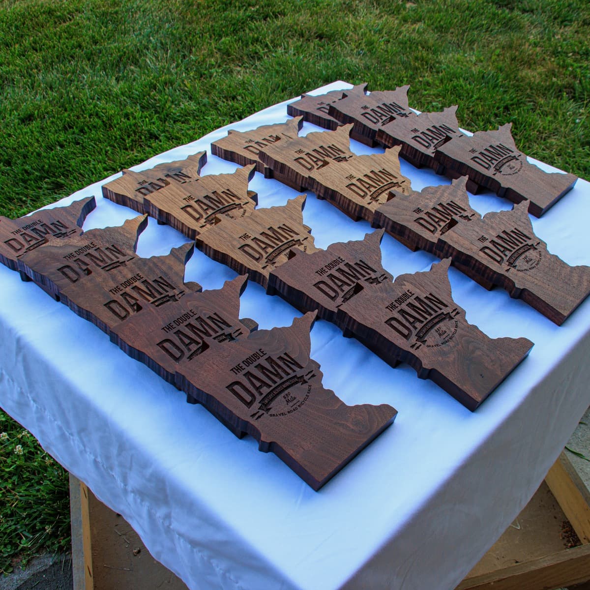 Array of twelve CNC-engraved wooden Minnesota-shaped trophies displaying "THE DOUBLE DAMn" text, arranged in rows on a white tablecloth outdoors, showing varying wood tones from light oak to dark walnut with consistent engraving quality across all pieces.