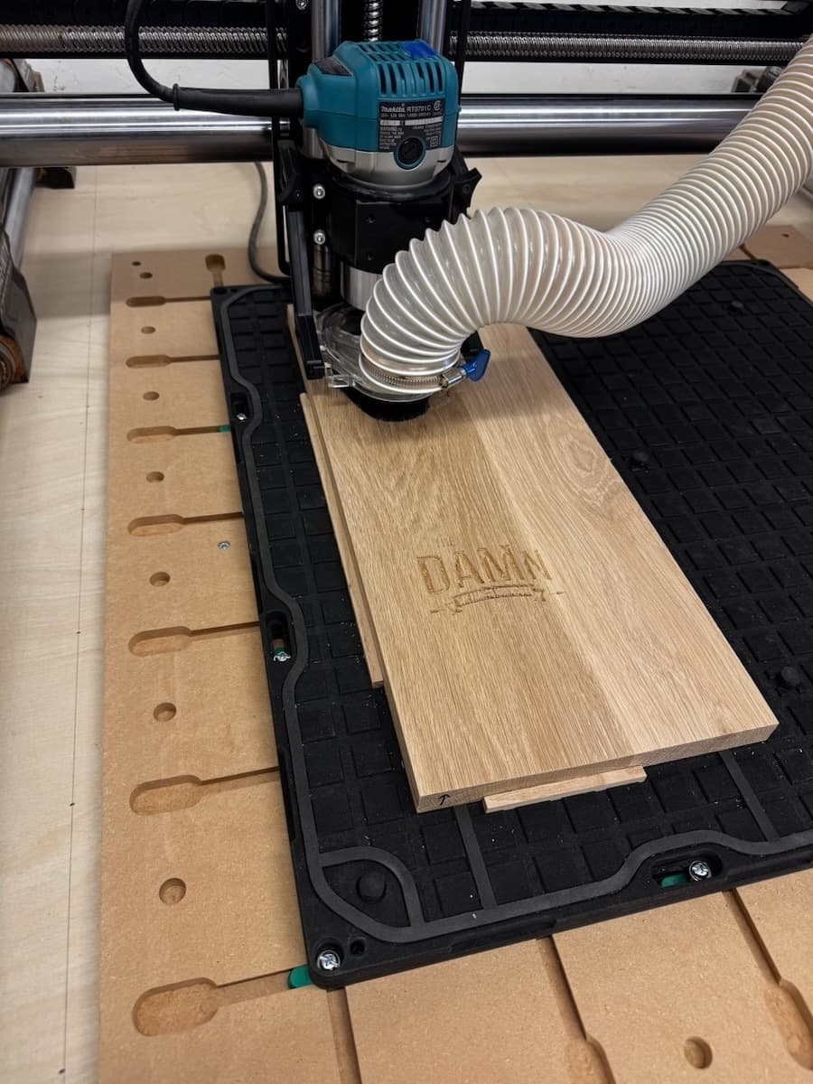 CNC router setup with white oak trophy plaque positioned on the machine bed, showing 'The DAMn' engraving in progress with the spindle positioned for cutting