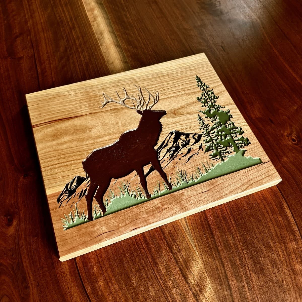 CNC-carved cherry wall art featuring a majestic elk with detailed antlers in silhouette, surrounded by mountains and evergreen trees with hand-painted green accents