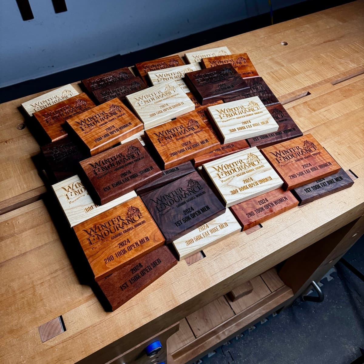 Overhead view of complete set of Winter Endurance 2024 trophy plaques arranged in rows showing all wood species variations—walnut, cherry, and ash—with CNC-engraved text for placement categories and race information